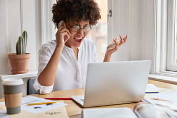 Outraged dark skinned entrepreneur has unpleasant talk with colleague via smart phone, tries to solve financial problem, looks at screen of laptop and gestures angrily, surronded with many papers