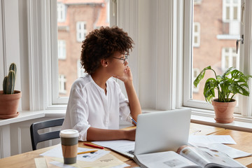 Concentrated young successful businesswoman with Afro hairstyle, checks financial documentations,...