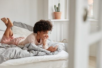 Cheerful teengae girl lies in bed, uses mobile phone, recieves text message from boyfriend, wears...