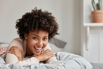 Candid shot of pleasant looking joyful black woman has toothy smile, curly dark hair, lies on stomache in bed, looks directly at camera with cheerful expression, spends morning in bedroom has lazy day