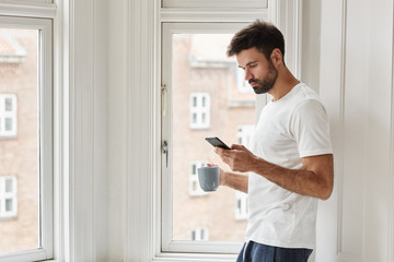 Handsome concetrated blogger holds modern mobile phone, installs application for creation new publication on personal website, dressed in domestic clothes, drinks aromatic coffee or cappuccino