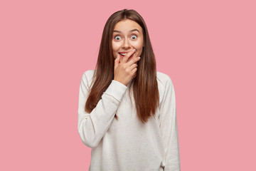 Surprised cheerful rejoiced teenager looks positively at camera, covers mouth, exclaims with astonishment, doesnt expect to recieve unexpected proposal from boyfriend, stands against pink wall