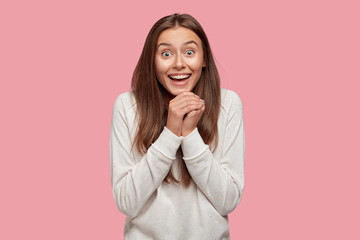 People, unexpectedness and emotions concept. Overjoyed European woman clasps hands from happiness, has broad smile and eyes full of delight, dressed in white sweatshirt, stands indoor alone.