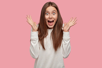 Amazed brunette young lady exclaims with happiness, clasps hands, has surprised happy expression, dressed in white oversized sweatshirt, stands against pink studio wall. People and emotions concept