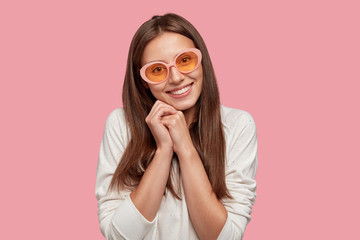 Attractive lovely girl with satisfied content expression, keeps hands together, wears fashionable...