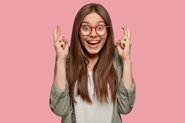 I wish good luck. Pretty youngster with happy expression keeps fingers crossed, believes in something wonderful, has delighted look, dressed in casual clothes, isolated over pink background.