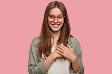 People, dedication and promise concept. Happy girlfriend keeps hands on heart, swears be loyal to boyfriend, expresses gratitude, wears spectacles, casual shirt, stands against pink studio wall. - obrazy, fototapety, plakaty