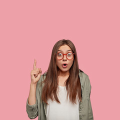 Disturbed youngster feels surprised, points with index finger upwards cant believe such sales exist, wears transparent glasses with red rim, isolated over pink studio wall. Omg, look above please!