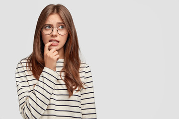 Photo of worried beautiful European woman bites fore finger and looks with hesitation aside, doubts...