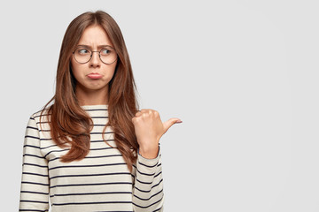 People and dislike concept. Displeased lovely woman purses lips, has offended expression, expresses antipathy, points with thumb left, stands against white blank studio wall for your advertisement