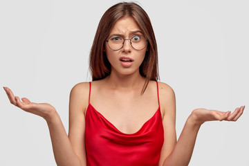 Fototapeta premium Headshot of indignant pretty young female stands in clueless pose, spread hands, cannot understand what is happening, wears spectacles and domestic clothes, stands against white wall. Whats problem?