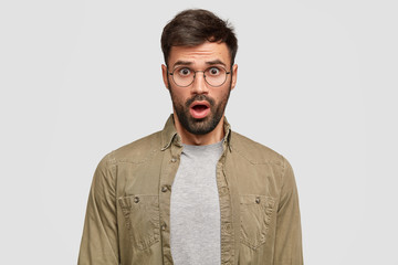 Photo of stunned emotive handsome guy has bewildered look at camera, opens mouth from surprisement, stands against white background in studio. People, emotions and facial expressions concept