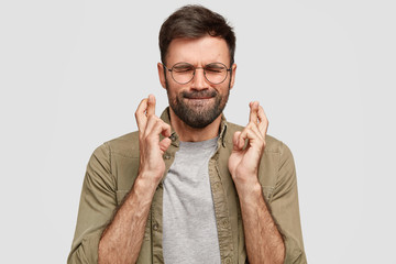 Portrait of handsome unshaven guy presses lips, closes eyes, keeps fingers crossed, has strong believe in good luck before important trip, wills everything would be fine. God, help me please!