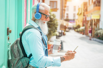 Trendy senior man using music smartphone app and drinking coffee in downtown center