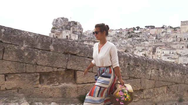 Beautiful elegant young woman walking in Matera Italy on a high balcony viewpoint with landscape. Fashion dress with hat and skirt.