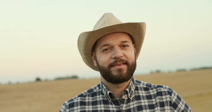 Portrait shot of the young farmer in a hat looking at the side and then smiling to the camera in the field. Close up.