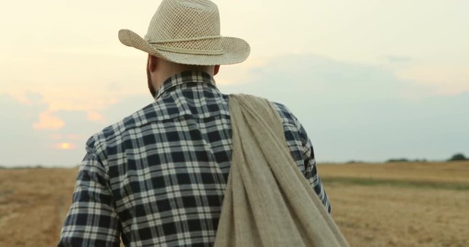 Rear of the young farmer man in a hat and plaid shirt and with a sack over the shoulder walking the field during the harvest season.