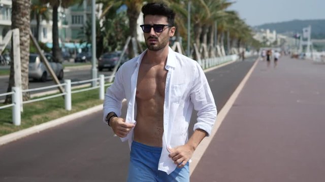 Attractive fit athletic young man soaking in the sun on seaside boardwalk or seafront, wearing white shirt open on naked torso, in Nice, France on the French Riviera