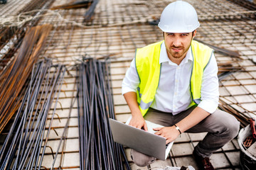 Industrial construction engineer working on laptop, wearing safety equipement and coordinating...