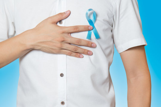 Men's healthcare concept - close up of male hand pointing to light blue ribbon for prostate cancer at left chest on blue background.
