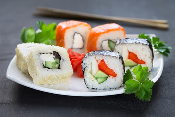 Sushi on a square white plate with parsley and chopsticks on a dark background.