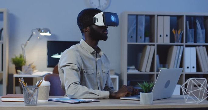 Young African American male office manager sitting late in the evening in hi office, having a VR headset, scrolling and taping in the air.