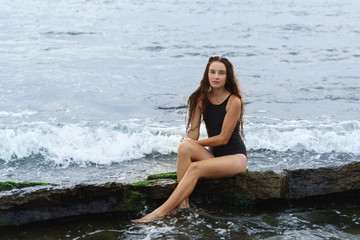 A beautiful slender long-haired girl in a black swimsuit sits on a stone on the sea