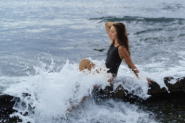 A happy beautiful long-haired girl in a black swimsuit sits on a rock in a foam of sea spray