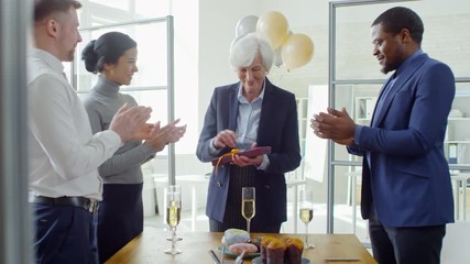 Handheld shot of cheerful senior woman opening present given to her by colleagues and saying thank you to them during retirement party in office