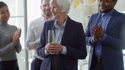 Tilt up handheld shot of smiling elderly woman holding glass of champagne at retirement party, her colleagues looking at her and clapping hands in the background