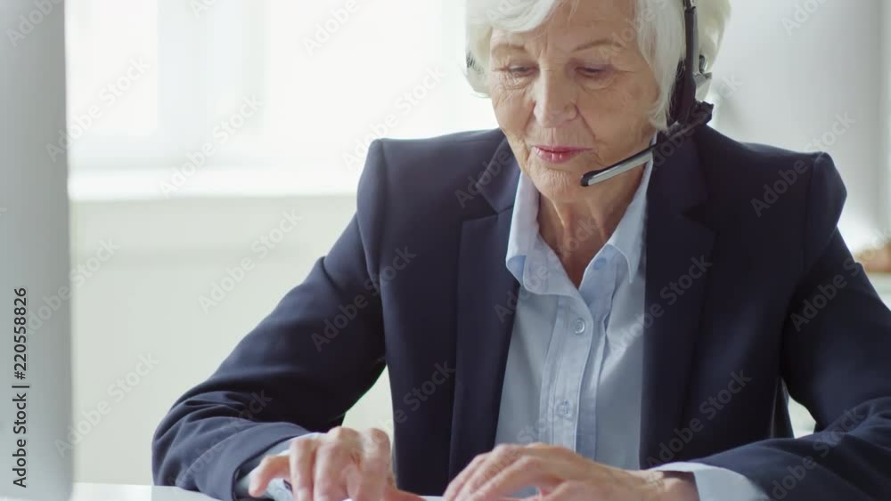 Poster medium shot of polite elderly woman using desktop computer and talking to client while working in ca