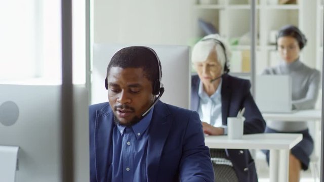 Tilt down shot of middle-aged black man in headset typing on desktop computer while communicating with client at work in call center