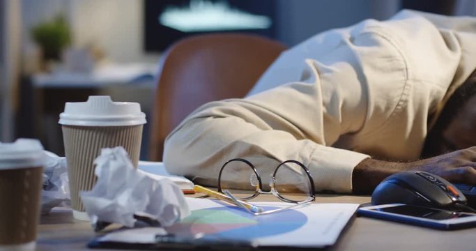 Close up of the exhausted African American man office worker sleeping on the desk full of empty coffee cups in the night.