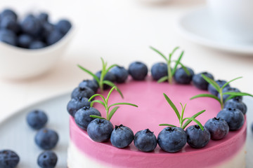 The two-color blueberry mini cheese cake. Round no bake cheesecake, bowl with blueberries, cup of coffee and bowl with sugar. Top of cake decorated by rosemary herbs. White table.