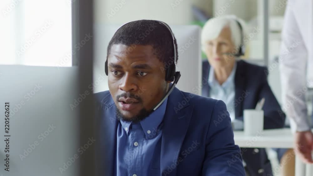 Poster tilt down shot of concentrated black man in suit and headset using desktop computer and talking to c