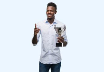 Young african american man holding trophy surprised with an idea or question pointing finger with...