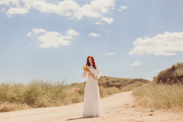 portrait of a beautiful red-haired girl in a white vintage wedding dress in full growth with a bouquet of reeds on the background of the road and nature