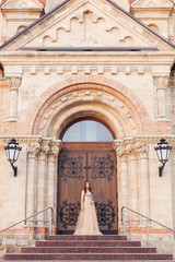 Obraz premium portrait of a young bride, a red-haired girl in a long dress of tulle and pajetok stands on the steps near wooden doors with a forged pattern of an ancient church, a castle with a place for text