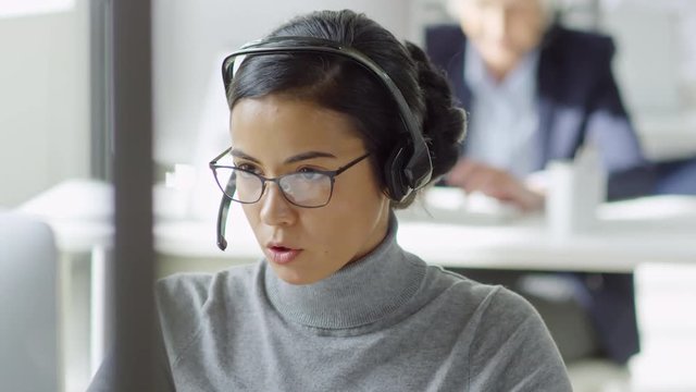 Medium shot of attractive young mixed race woman in glasses helping customer using desktop computer while working in call center