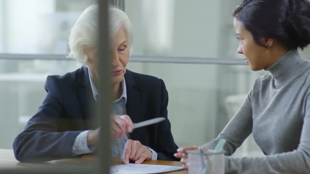 Medium Shot Of Two Diverse Female Office Workers Sitting At Desk And Having Conversation About Work