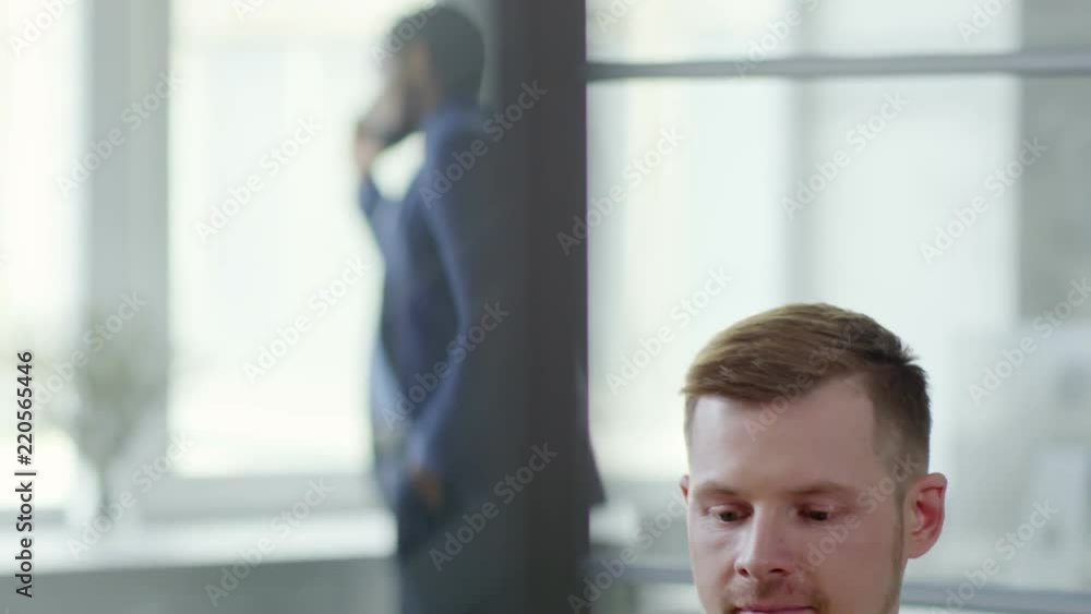 Poster Tilt down shot of optimistic Caucasian young man smiling at camera while working with laptop computer in office, his black colleague having telephone call in the background
