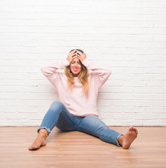 Young adult woman sitting on the floor over white brick wall suffering from headache desperate and stressed because pain and migraine. Hands on head.