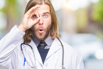 Young handsome doctor man with long hair over isolated background doing ok gesture shocked with surprised face, eye looking through fingers. Unbelieving expression.