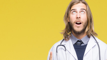 Young handsome doctor man with long hair over isolated background amazed and surprised looking up and pointing with fingers and raised arms.