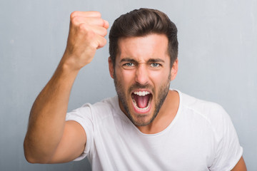 Handsome young man over grey grunge wall annoyed and frustrated shouting with anger, crazy and yelling with raised hand, anger concept