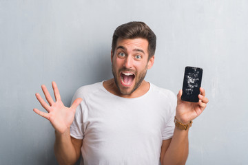 Handsome young man over grey grunge wall showing broken smartphone screen very happy and excited,...