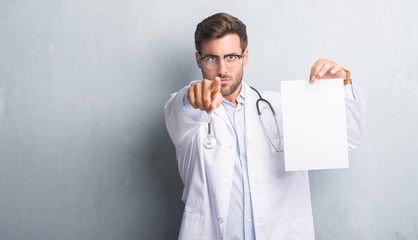 Handsome young doctor man over grey grunge wall holding blank sheet contract pointing with finger to the camera and to you, hand sign, positive and confident gesture from the front