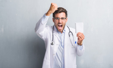 Handsome young doctor man over grey grunge wall holding blank notebook annoyed and frustrated shouting with anger, crazy and yelling with raised hand, anger concept