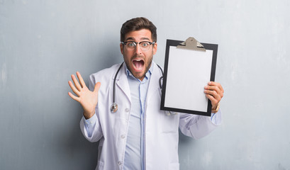 Handsome young doctor man over grey grunge wall holding clipboard very happy and excited, winner expression celebrating victory screaming with big smile and raised hands