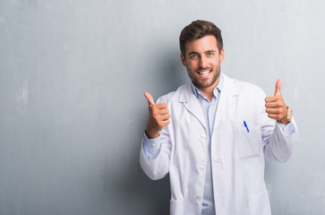 Handsome young professional man over grey grunge wall wearing white coat success sign doing positive gesture with hand, thumbs up smiling and happy. Looking at the camera with cheerful expression